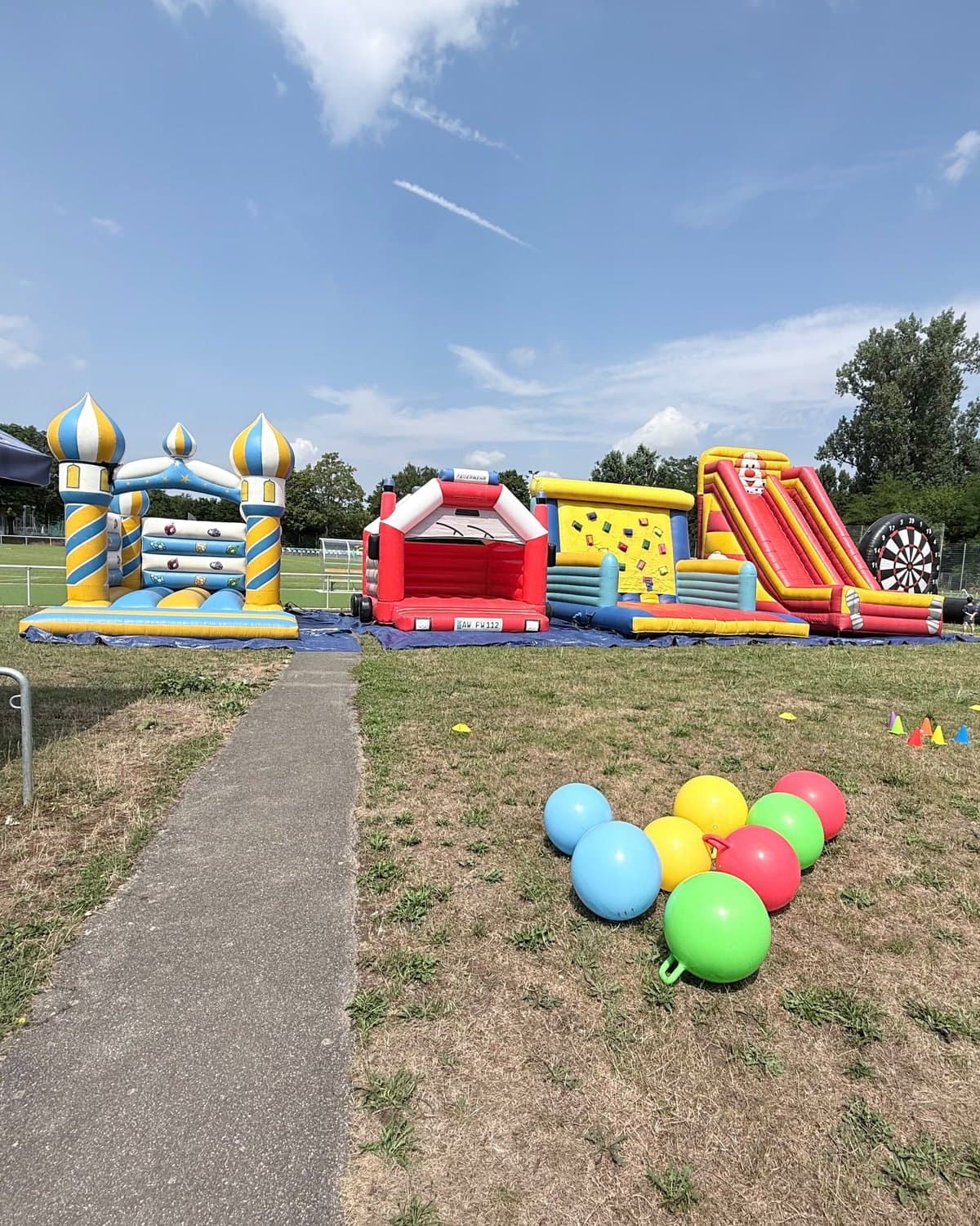 Aufgebaute Hüpfburgen, Kletterwand, Riesenrutsche und Riesendart auf einer Sportplatzwiese, bunte Hüpfbälle im Gras