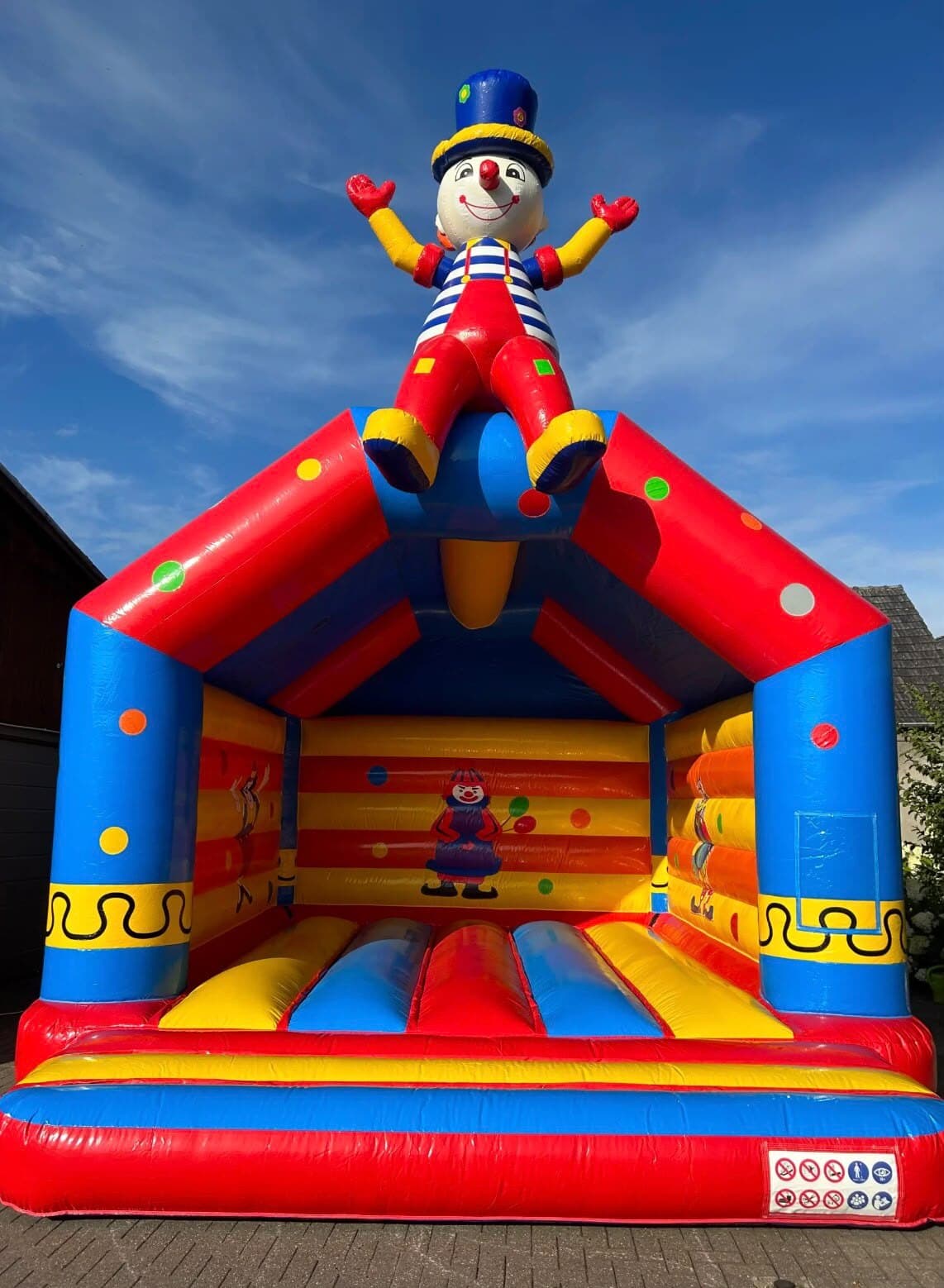 Hüpfburg mit großer Clown-Figur mit Zylinder auf dem Dach, rot, blau und gelb mit bunten Tupfen, vor blauem Himmel